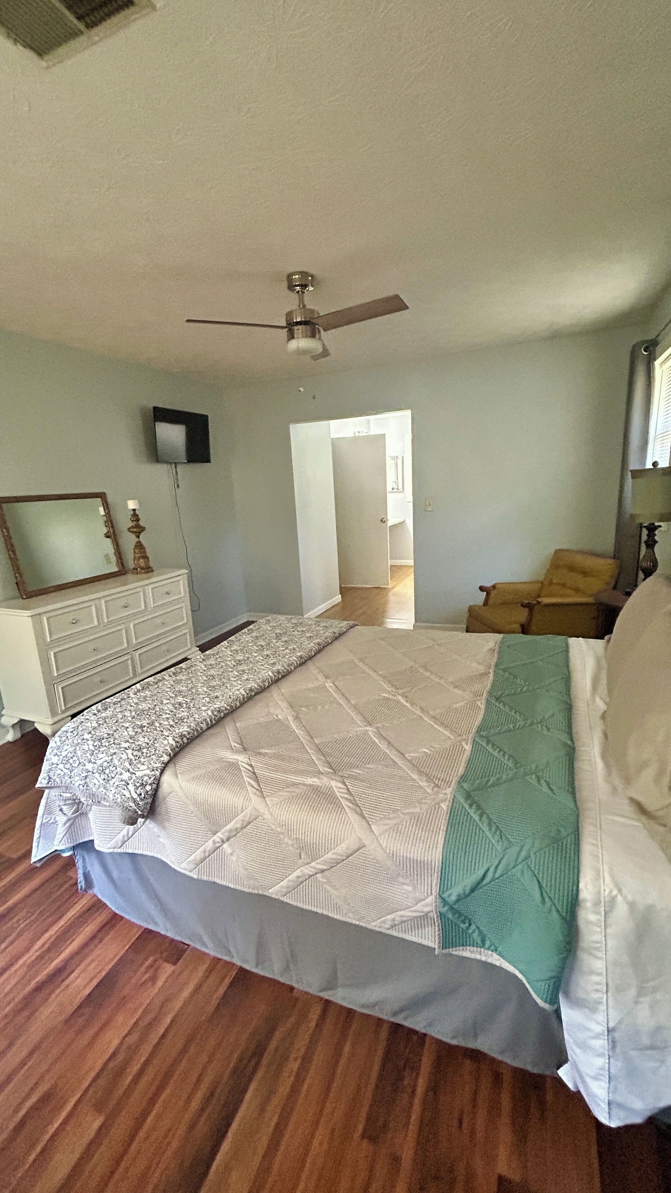 Une chambre à coucher comprenant un lit, une commode, un ventilateur de plafond et une porte donnant sur une autre pièce.
