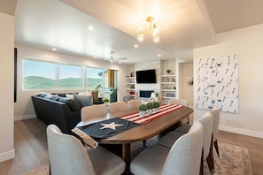 A modern living room with a large window, a dining area with a wooden table and chairs, a ceiling fan, and a mounted TV. - DVE Adventure Basecamp | Saltwater Hot Tub | Ski Shuttle to Slopes (Heber City)