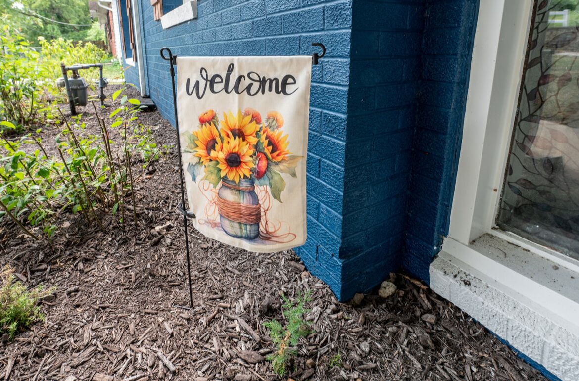 A welcome banner with sunflowers and a basket, hanging on a blue house exterior.