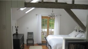 Ein Schlafzimmer mit Holzboden, einem Bett mit weißer Bettwäsche, einem Nachttisch, einem Bücherregal, einem Schreibtisch mit Lampe, einem Stuhl und einem Fenster mit Vorhängen.