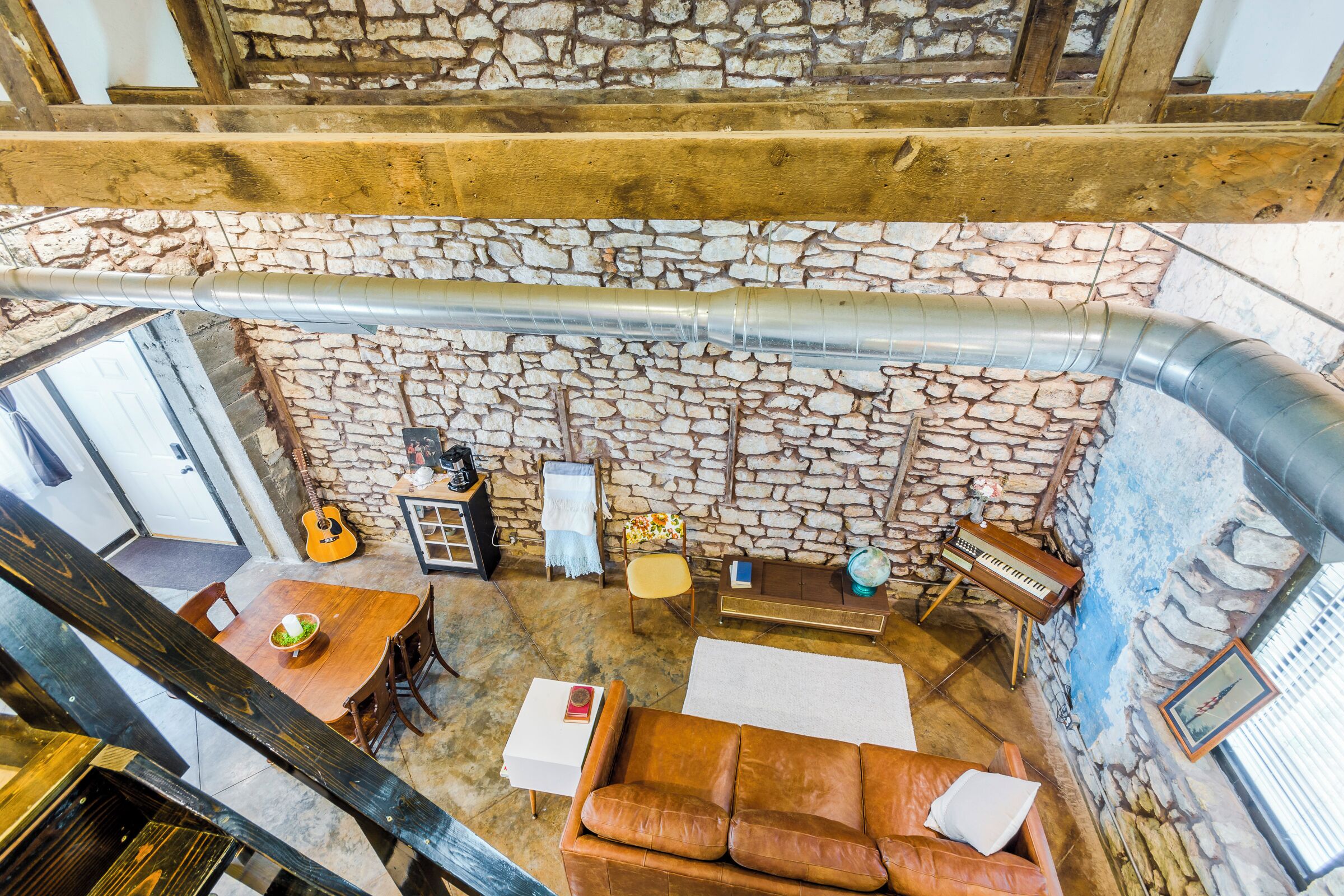 A rustic room with stone walls, a wooden ceiling, a brown leather sofa, a dining table, and a guitar.