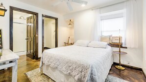 Une chambre à coucher comprenant un lit, un ventilateur de plafond, une fenêtre avec des rideaux, une table de chevet et une chaise.