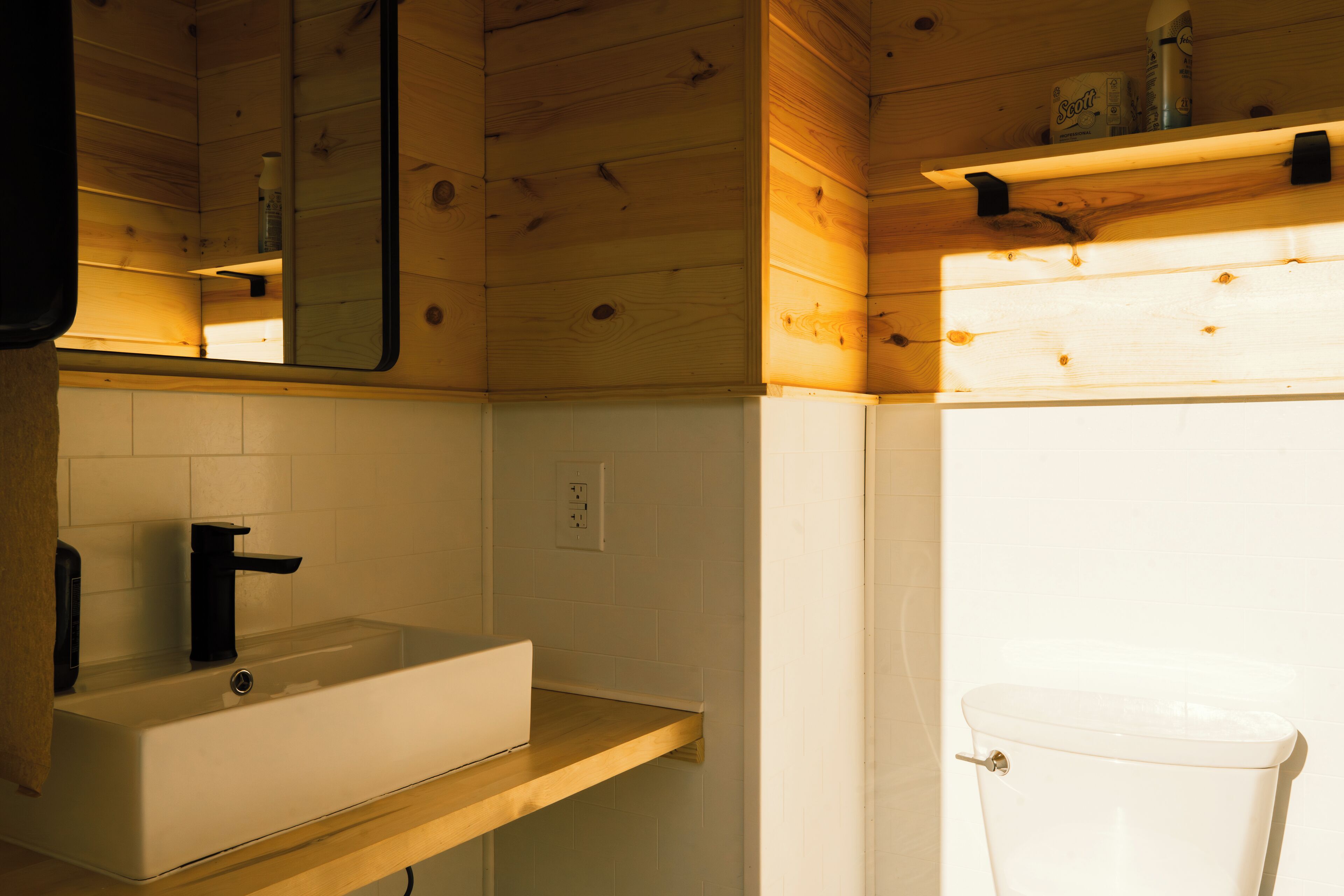 A bathroom with a wooden interior, a white sink with a black faucet, a mirror above the sink, and a toilet visible to the right.