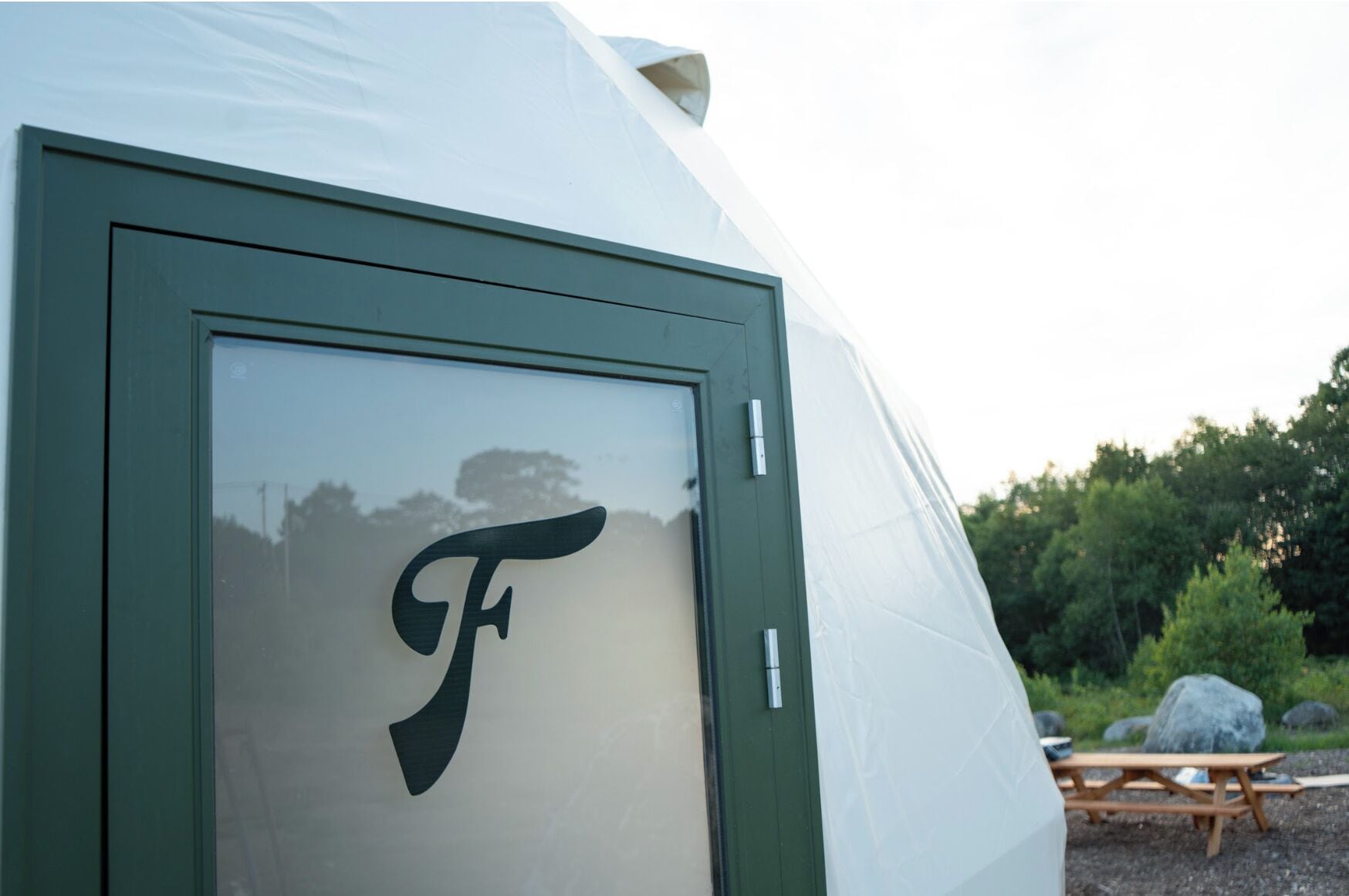 Une tente ornée d’un grand logo en forme de « F » sur la porte, entourée d’un paysage naturel et agrémentée d’une table de pique-nique.