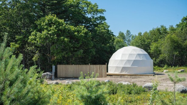 A geodesic dome structure in a natural setting with a wooden fence and dense greenery. - Ferncrest Acadia (Sedgwick)