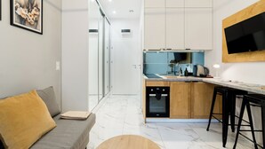 A modern kitchen with a built-in oven, a dining area with a wooden table and bar stools, and a living area with a sofa and a painting on the wall.