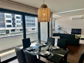 A modern dining area with a glass table set for four, a black sofa, a television, and a city view through large windows.