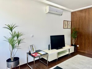 A modern living room with a flat-screen TV on a white TV stand, two potted plants, a wooden door, and a wall-mounted air conditioner.