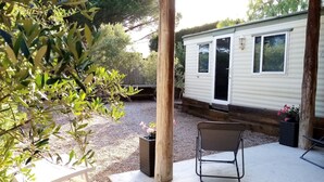 A covered patio area with a white table and a metal chair, a gravel path, and a mobile home in the background. - Mobile home in Ramatuelle - plaine de Pampelonne (Ramatuelle)