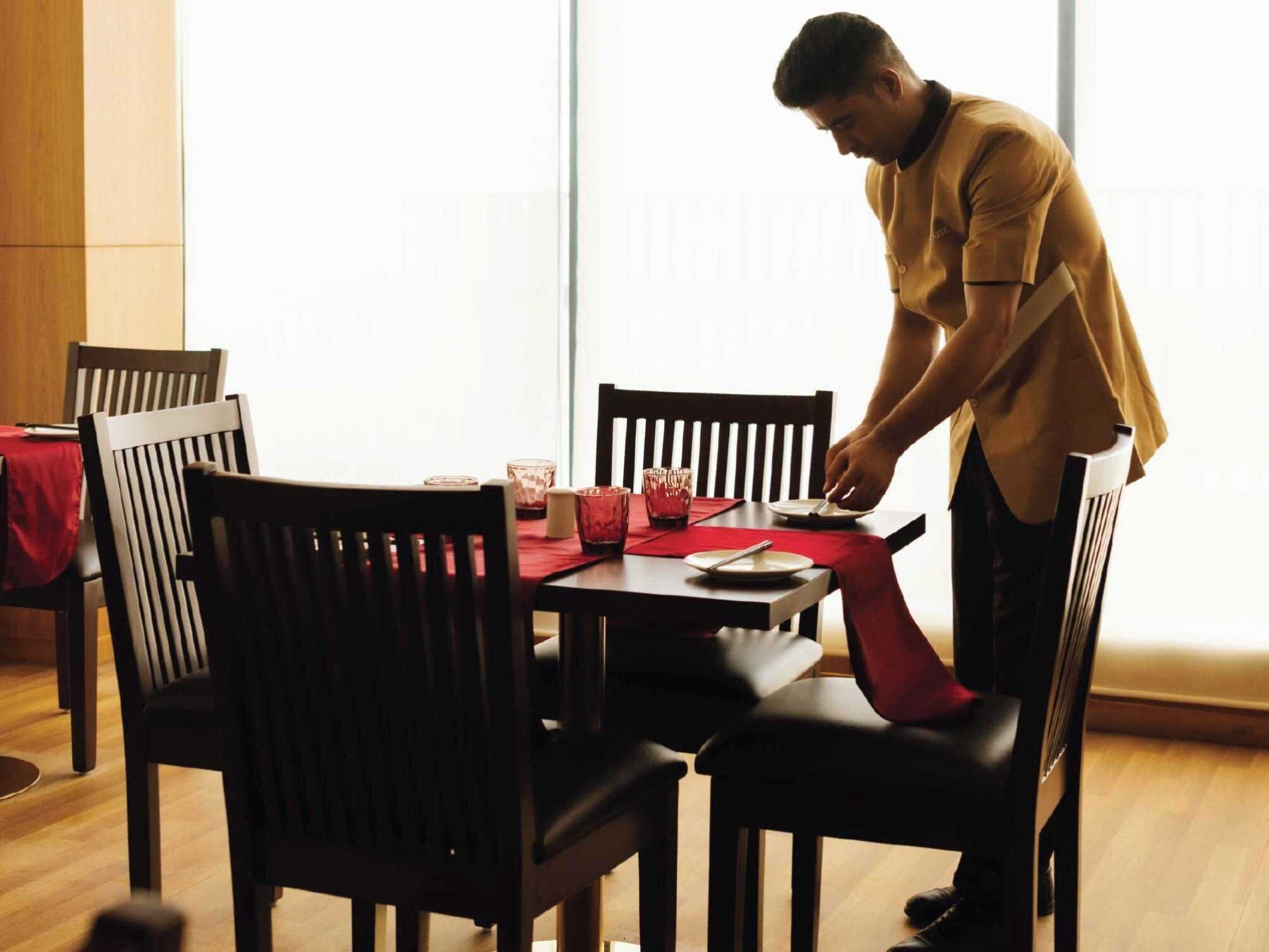 A dining area with a table set for two, a person serving food, and a window providing natural light.