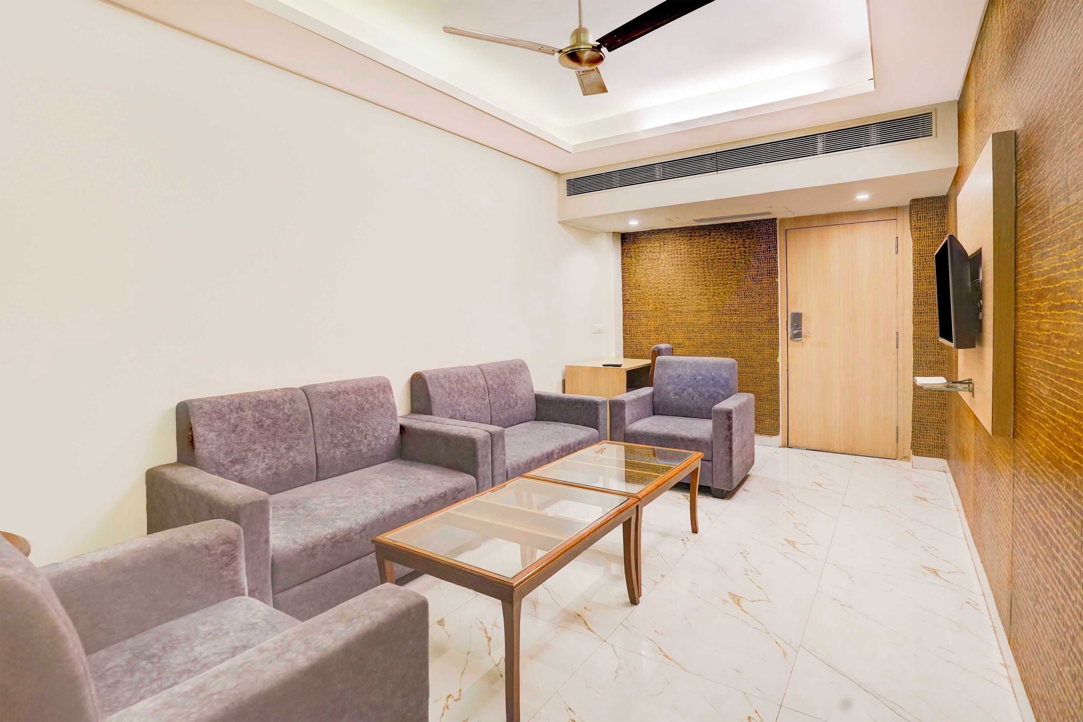 A modern living room with a sofa set, a glass-top coffee table, a ceiling fan, a TV mounted on the wall, and a wooden door.