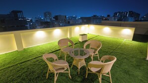 An outdoor patio with a round table and wicker chairs, surrounded by artificial grass, with city buildings in the background. - Anand's Abode By Yellow Bells (Hyderabad)