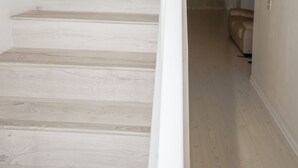 Un escalier aux murs blancs et au plancher de couleur claire mène à une pièce où se trouvent un lit et une fenêtre.