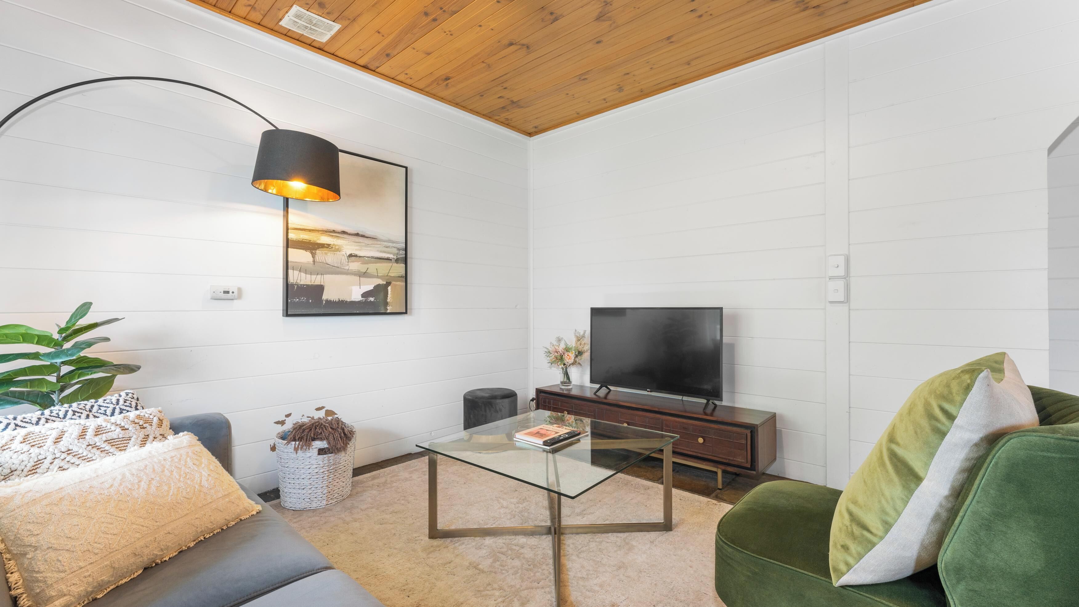 A modern living room with a glass coffee table, a green sofa, a grey couch, a black TV stand, and a framed picture on the wall.