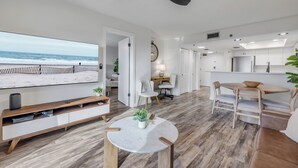 Ein modernes Wohnzimmer mit großem Fernseher, Holzboden, Deckenventilator und Blick auf den Strand.