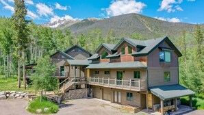 A multi-story log cabin with a balcony, set in a forested area with mountains in the background.