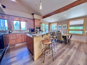 A modern kitchen with wooden cabinets, a central island, bar stools, and a dining area with a table and chairs.