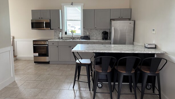 A modern kitchen with grey cabinets, a stainless steel refrigerator, a built-in oven, and a marble countertop. There are four black chairs around the island.