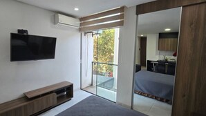 Un dormitorio moderno con una televisión de pantalla plana, una mesita de noche de madera y un espejo grande que refleja un balcón con vistas a árboles.