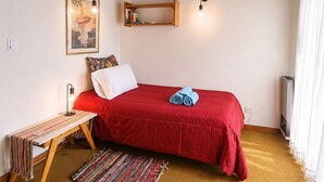 Une chambre avec un lit rouge, un banc en bois, un tapis coloré, une étagère fixée au mur et un tableau encadré.