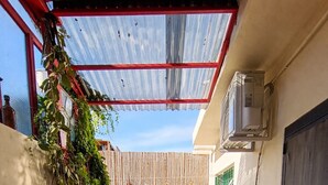 Un étroit couloir extérieur avec un hamac, des plantes en pot, un sol carrelé et une porte sur la droite.