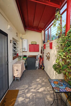 A small, enclosed patio area with a red ceiling, a tiled floor, a table with a checkered tablecloth, a water heater, and a wall-mounted shelf with various items.