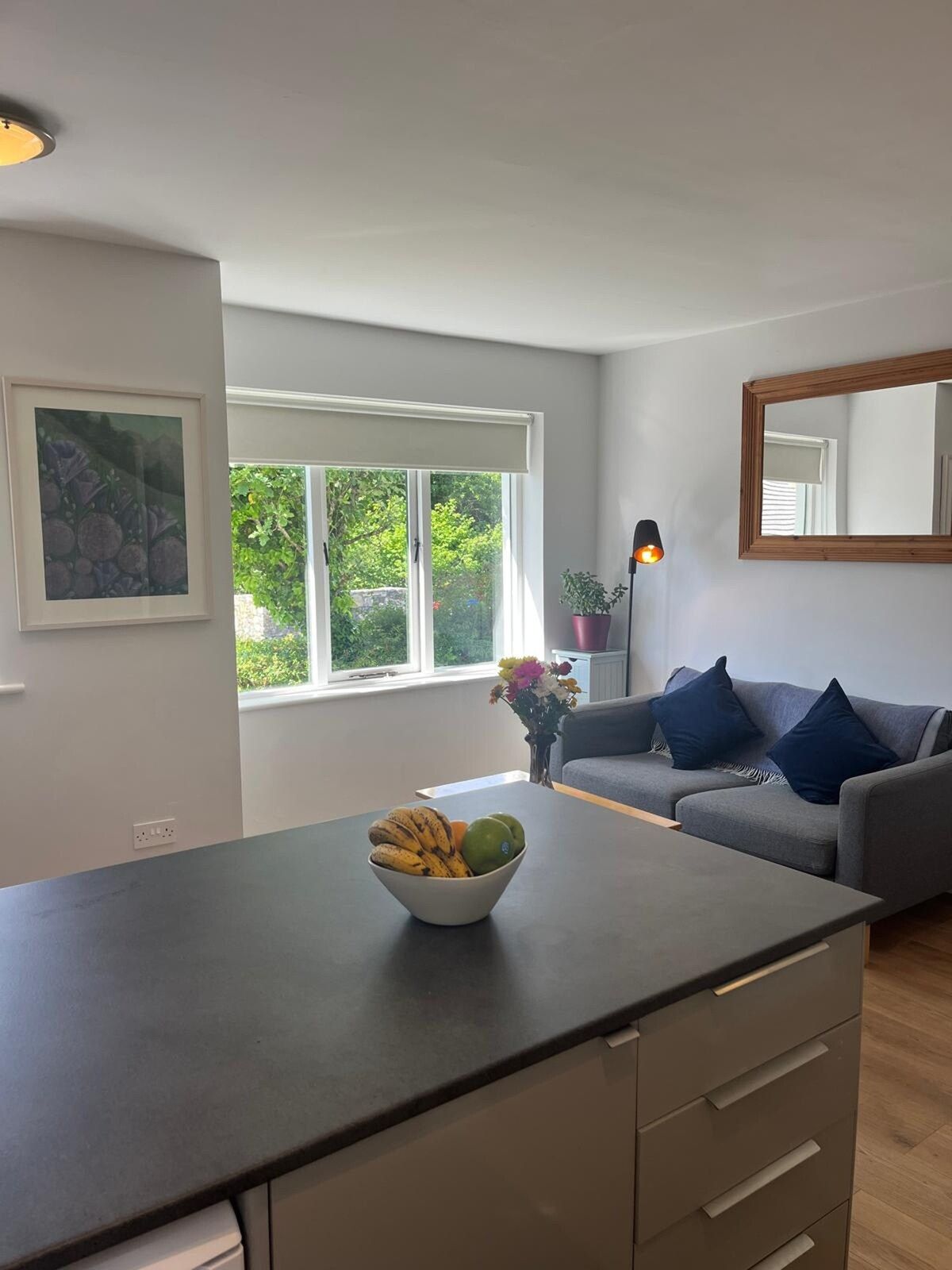 Un salon moderne avec un canapé gris, une table à manger sur laquelle se trouve un saladier de fruits, une grande fenêtre donnant sur un paysage verdoyant et un lampadaire.
