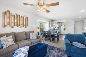 A modern living room with a sofa, ottoman, and blue armchair. There is a ceiling fan, a dining area with a table and chairs, and a kitchen in the background. - New beautiful condo 2-king bedrooms 2-full baths sleeps 6-7 next to SDC (Branson)