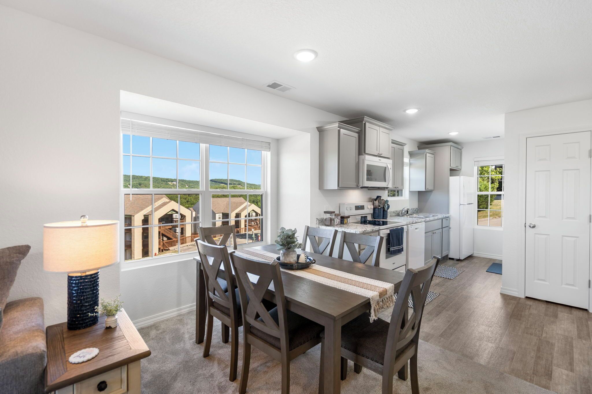 A modern kitchen with white cabinets, a dining table with chairs, and a view of a house through a large window.