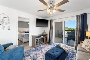 A modern living room with a fireplace, a ceiling fan, a TV mounted on the wall, and a view through the glass door to the outside. - New beautiful condo 2-king bedrooms 2-full baths sleeps 6-7 next to SDC (Branson)