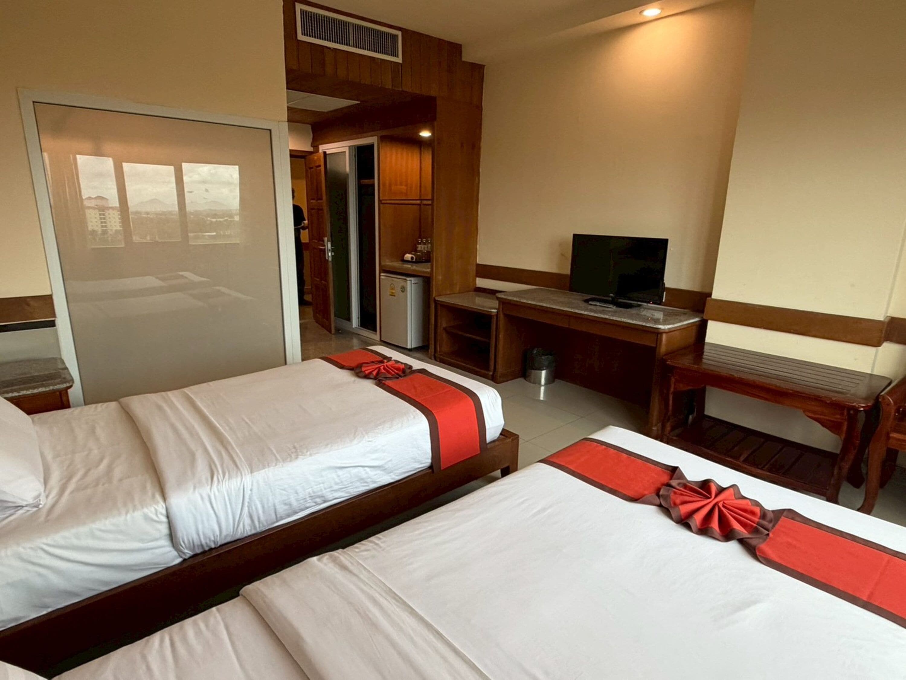 A hotel room with two beds, a desk, a television, and a small kitchenette.