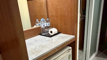 A compact kitchenette with a small refrigerator, a countertop, and a glass shelf holding water bottles and a roll of paper towels.