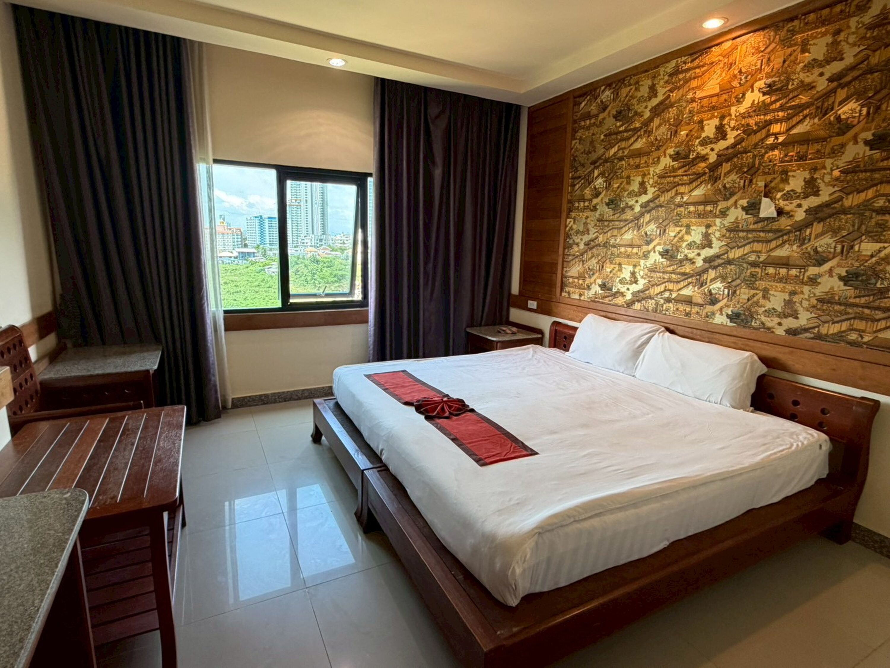 A hotel room with a large bed, a wooden headboard, a bench, a small table, and a view of buildings through a window.