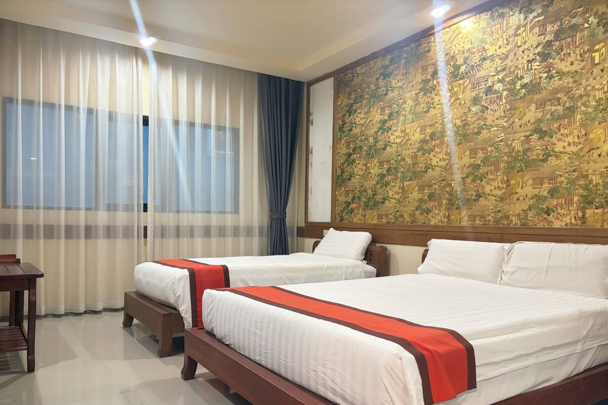 Une chambre d’hôtel avec deux lits, une table de chevet en bois, un bureau et une grande tapisserie au mur.