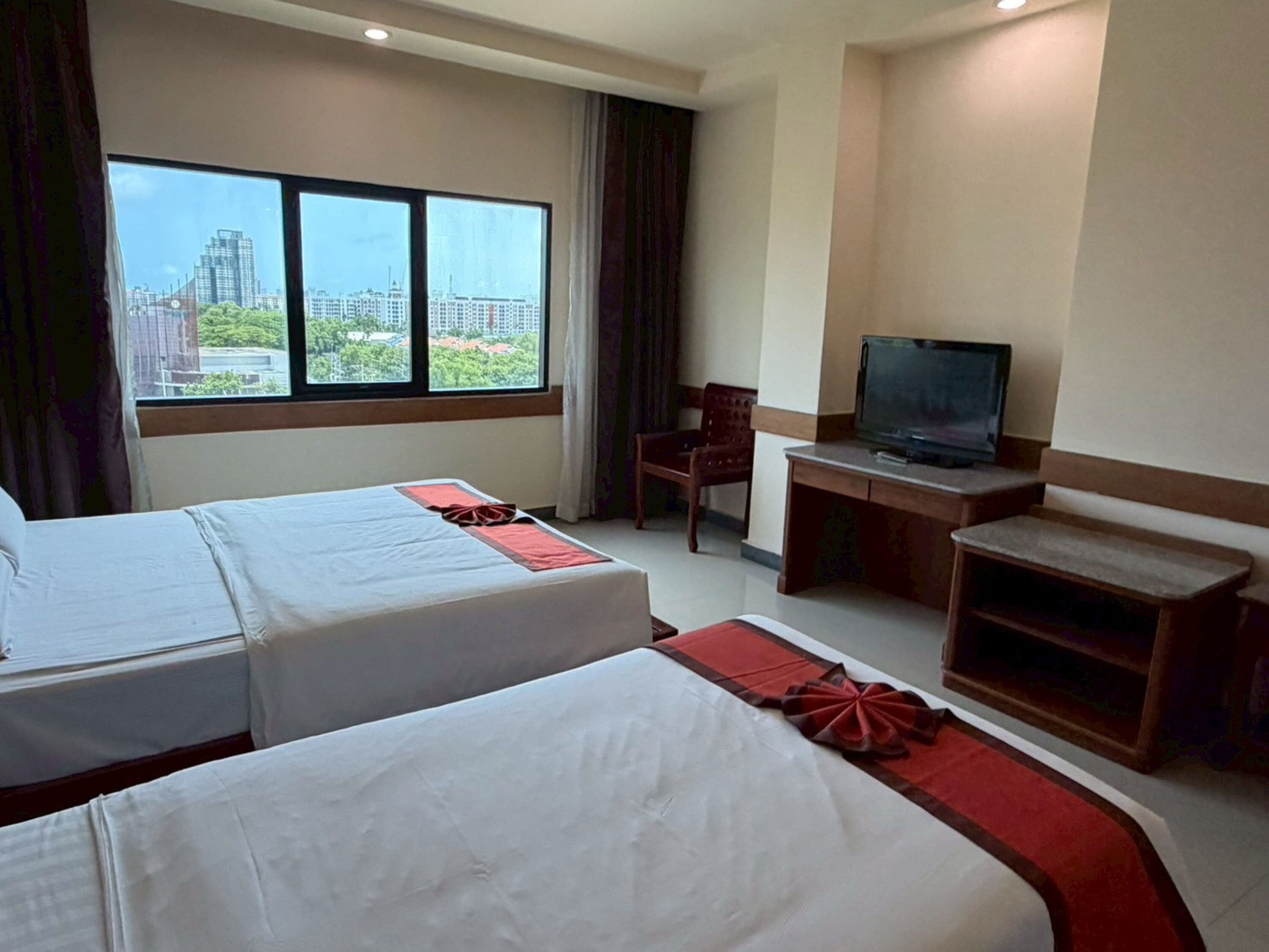 A hotel room with two beds, a desk with a television, and a view of a cityscape through large windows.