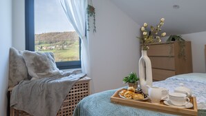 A bedroom with a view of a green landscape, a bed with a floral bedspread, a bedside table with a tray of pastries, a vase with flowers, and a wooden dresser. - River House Val Gorski kotar (Pribanjci)