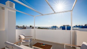 Un balcon ensoleillé, agrémenté de meubles blancs, d’une table en bois et offrant une vue dégagée sur la mer et les bâtiments qui se dressent à l’arrière-plan.