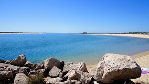 Une plage avec une eau d’un bleu limpide, un rivage sablonneux et de gros rochers au premier plan.