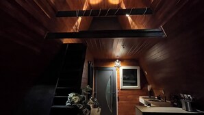 Ein Dachzimmer mit Holzwänden und schragem Dach. Eine Treppe führt zur oberen Etage, es gibt eine kleine Küchenzeile mit Spüle und Arbeitsfläche sowie eine Tür mit Fenster.