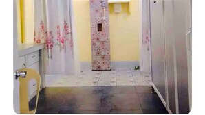 A narrow hallway with a tiled floor, a door with a floral-patterned door mat, and a yellow wall with a window.