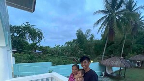 A man and two children on a balcony with a view of palm trees and a thatched structure.