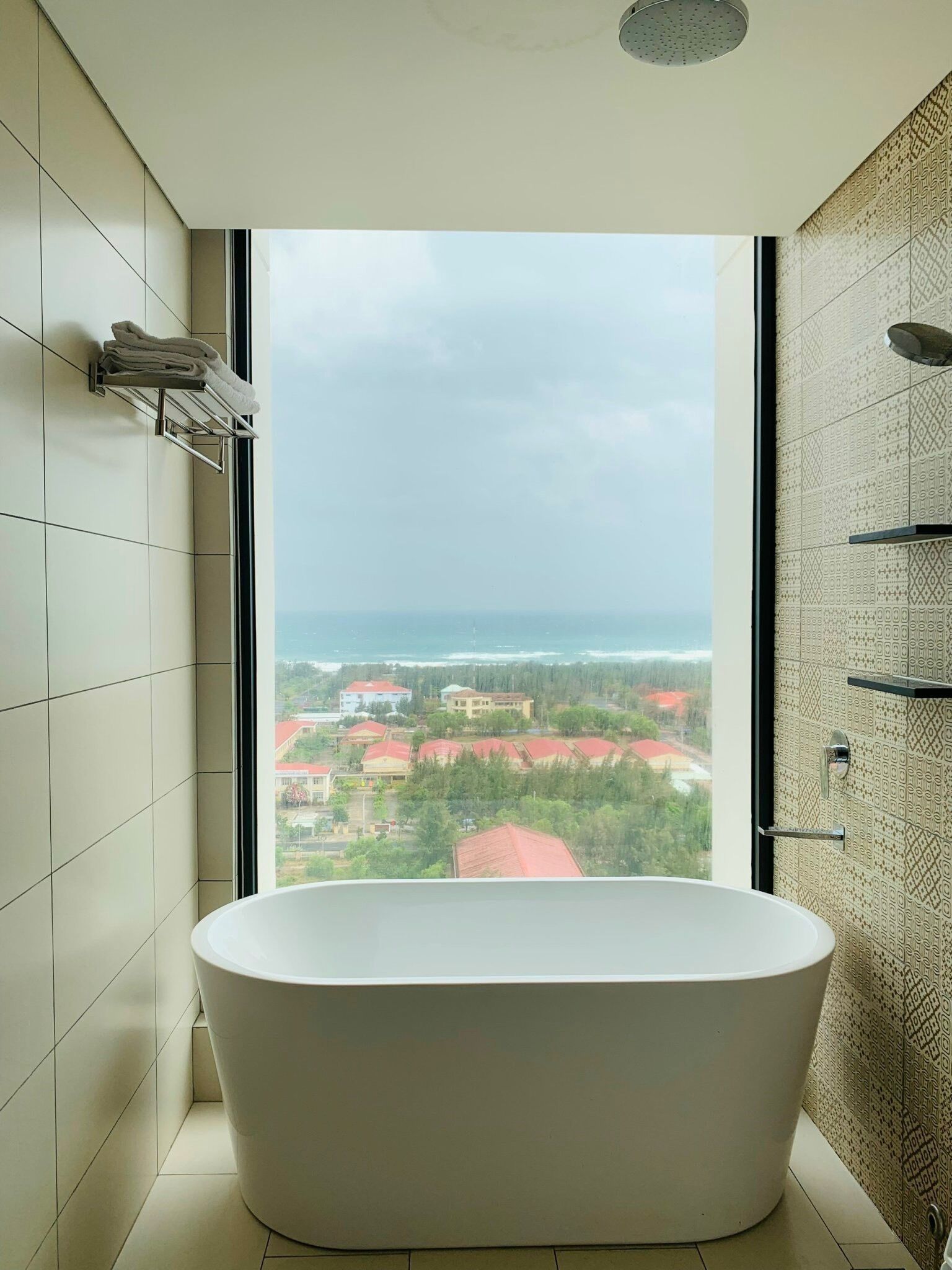 A modern bathroom with a freestanding bathtub, a large window with a view of buildings and greenery, and patterned tiles on the walls.