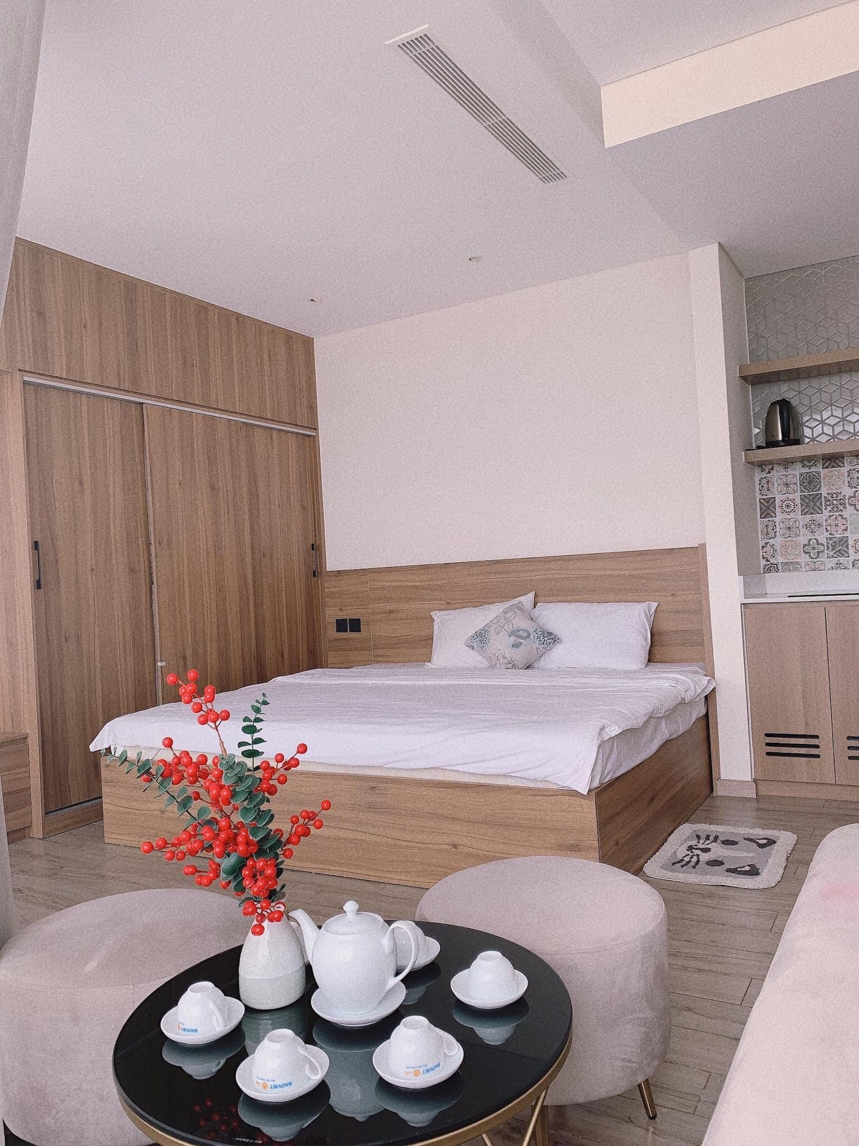 A modern bedroom with a wooden bed, white bedding, a small round table with tea set, and a decorative plant.