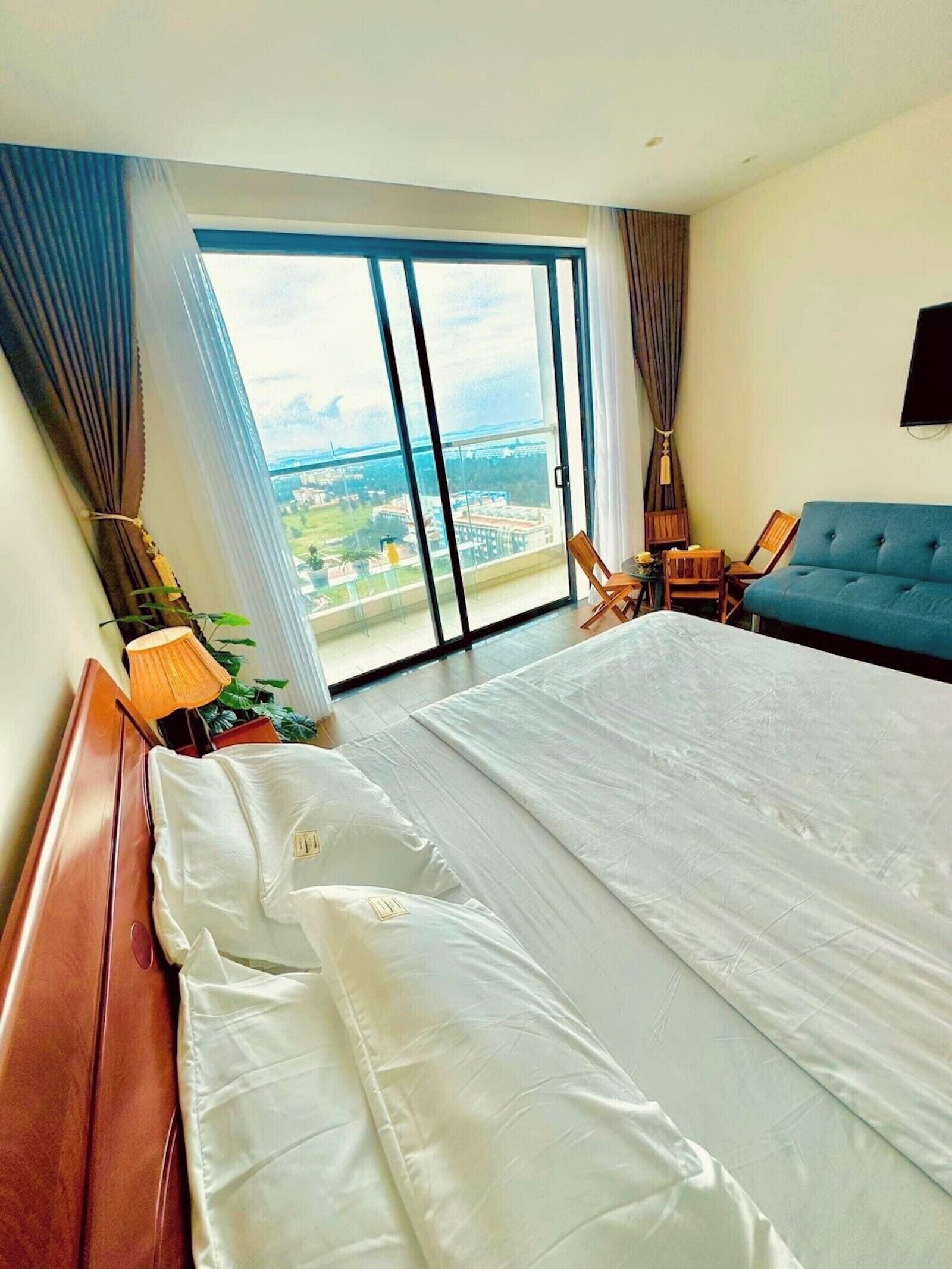 A hotel room with a large bed, two chairs, a sofa, a TV, and a view of a cityscape through a large window.