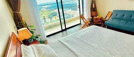 A hotel room with a large bed, two chairs, a sofa, a TV, and a view of a cityscape through a large window.