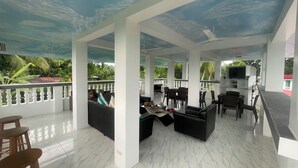 A covered outdoor seating area with white pillars, a TV, and a view of greenery.