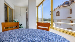 Une chambre avec une grande fenêtre, un lit recouvert d’une couvre-lit à motifs, une tête de lit en bois et une vue sur un immeuble doté d’un balcon.