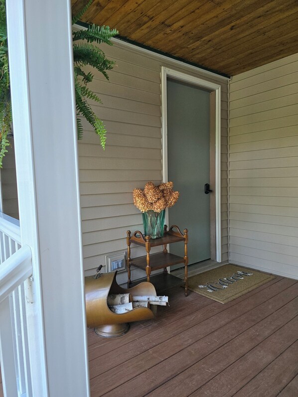 Un porche de madera con una puerta, un felpudo y una silla con un jarrón de flores.