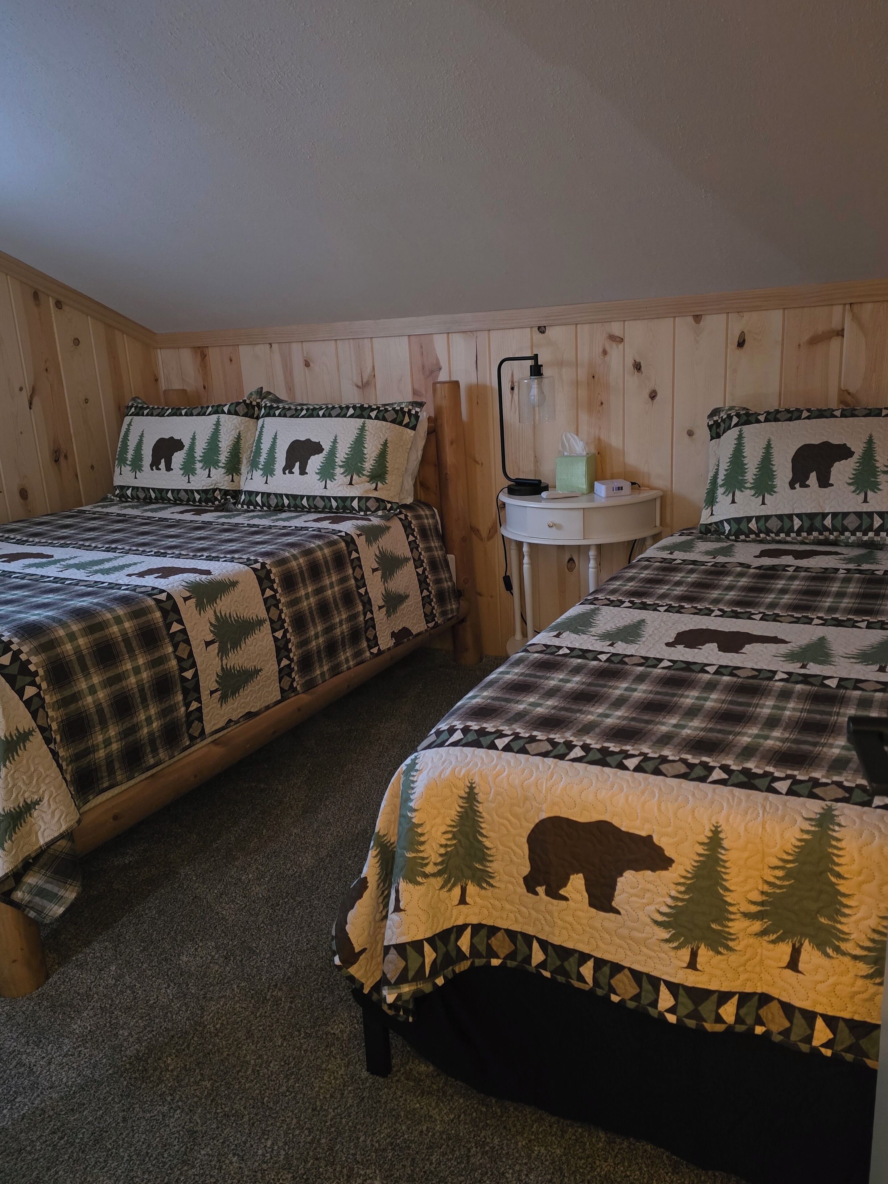 Two twin beds with forest-themed bedding, a small bedside table with a lamp, and a wall-mounted shelf.
