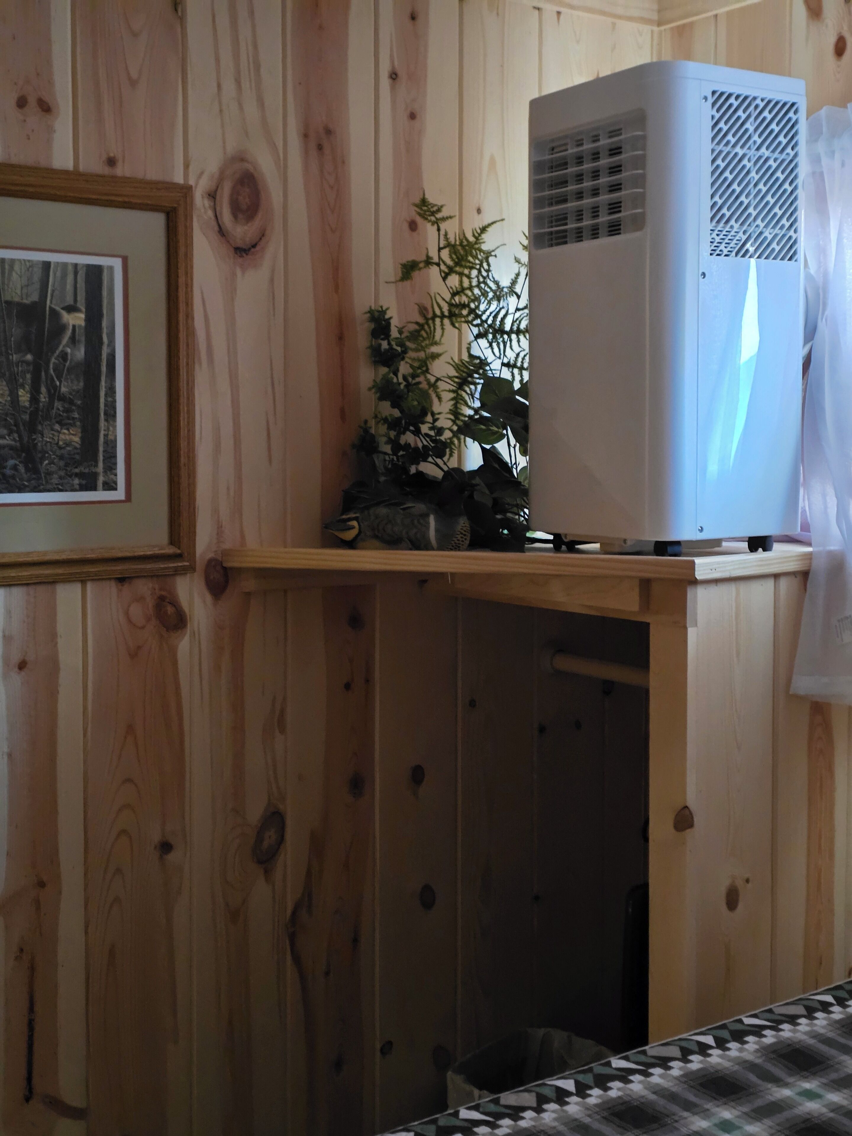 Una habitación con paredes de madera, un cuadro enmarcado, un aire acondicionado blanco y una planta pequeña en una estantería.